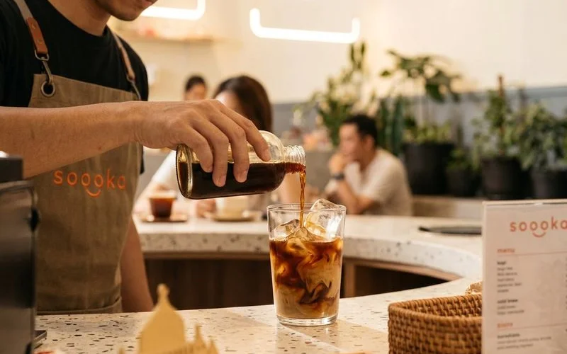 Barista at Soooka Cafe pouring cold brew kopi concentrate over ice in a clear glass with warm cafe background