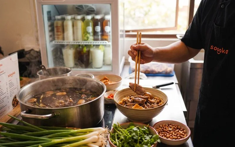 Chef preparing braised duck noodles at Soooka Cafe kitchen with fresh ingredients and steaming broth