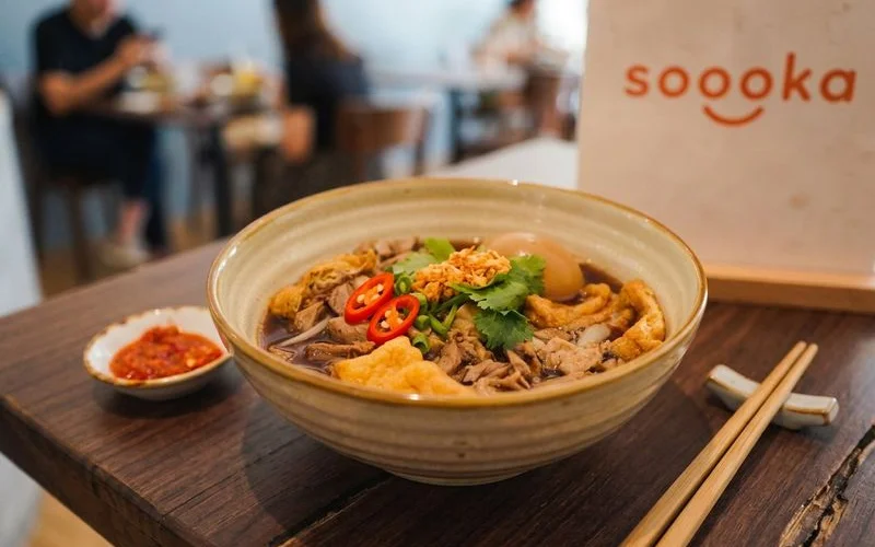 Complete bowl of Soooka braised duck flat white noodles garnished with fresh herbs and served with chilli sauce